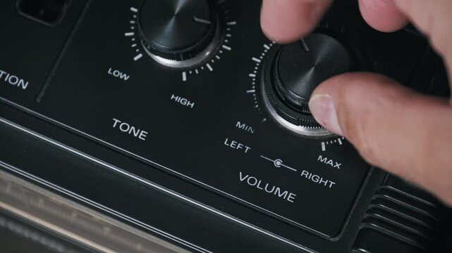 Angled macro view of fingers turning a plastic volume knob on a vintage boombox adjusting sound from low to high White dial markings and tone controls are visible on the black electronic panel