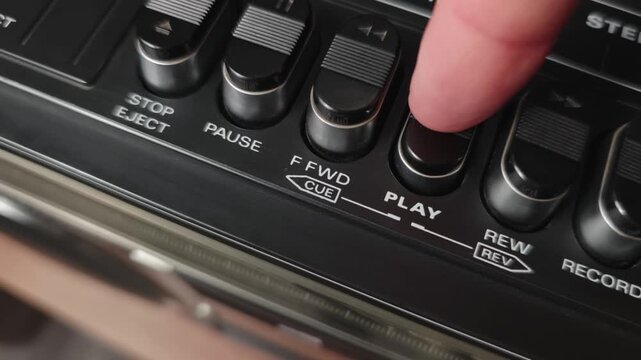 Angled macro view of a finger pushing the play button on a vintage black tape deck control panel showing ridged keys chrome accents and a slight pan across the fast forward and rewind controls