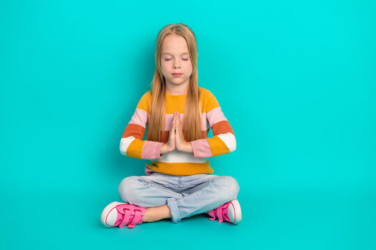 Young girl meditating in prayer pose on teal background wearing colorful sweater and sneakers, promoting relaxation concepts