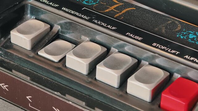 Macro of a finger pressing play and stop buttons on a vintage cassette player, showing dusty gray keys, German text labels, and worn chrome trim. Captures direct analog technology interaction.