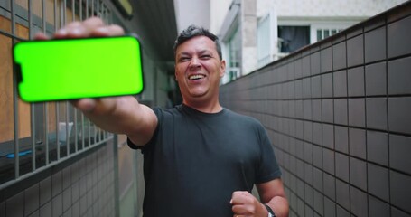 Man cheering with excitement and celebrating victory, holding smartphone and showing customizable green screen to the camera, urban corridor background, moment of triumph