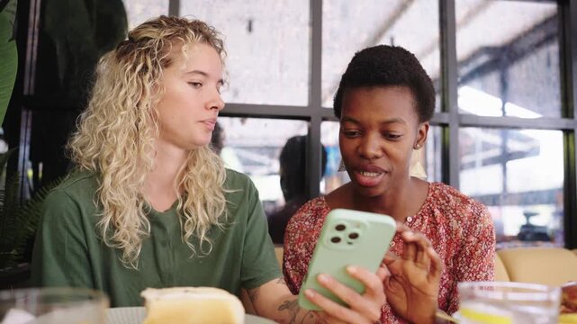 Two cheerful diverse women using smartphone and laughing in a cafe