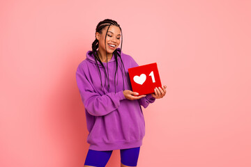 Smiling young woman holding a red box with a heart symbol against a pink background in casual sportswear