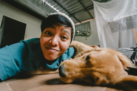 A real Asian man takes a selfie with a year-old golden retriever. The dog is licking its owner's face. Smiling People and fun playing together looking happily at the camera at home Pet and person - Powered by Adobe