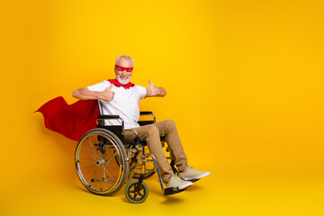 Funny grandpa superhero in wheelchair with red cape and mask smiling on bright yellow background promoting accessibility inclusion and rehab spirit