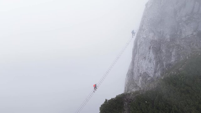 Alpinists climbing a long ladder on a sheer mountain cliff in dense fog, Dachstein, Austria