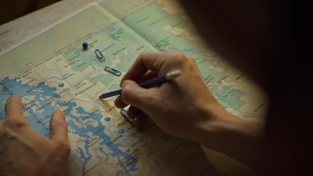 A person examines a detailed map while using a pen to take notes and paper clips to mark locations. The atmosphere feels warm and focused, perfect for trip preparation on a leisurely day