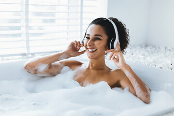 In a bright bathroom, a woman relaxes in a bubble-filled tub while wearing headphones. She smiles and enjoys her private time, surrounded by soft light and calm atmosphere.