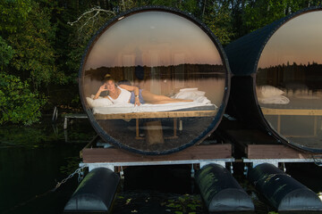 A woman enjoys a tranquil glamping getaway, relaxing in a floating barrel cabin with a panoramic window on an Estonian lake.