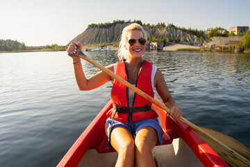 A happy smiling woman in a life jacket enjoys a summer adventure, paddling a red canoe at Rummu quarry, Estonia.