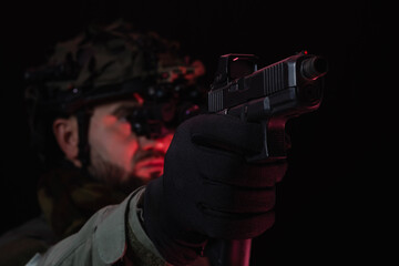 Special operations soldier in tactical gloves and night vision aims a modern semi-automatic pistol with a red dot sight.