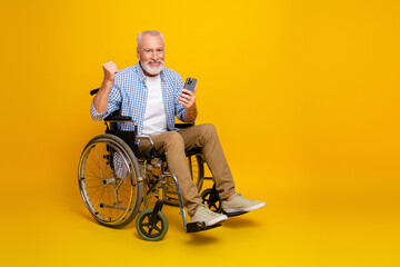 Cheerful grandpa in wheelchair celebrates success with a phone against a bright yellow background...