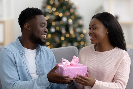 African American man gives woman Christmas gift, both smile with joy, celebrating festive holiday season.