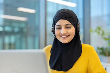 Muslim woman in black hijab wearing a headset, smiling at her laptop while providing friendly customer support in a modern office, professional and approachable agent