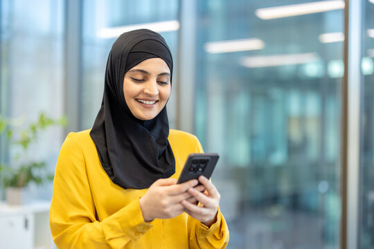 Young professional muslim woman wearing a hijab and yellow shirt, happily focusing on her smartphone screen while connecting, communicating, and working in a modern office environment