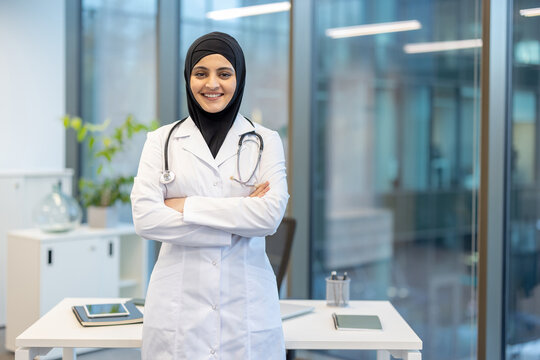 Fototapeta Muslim woman doctor wearing a hijab and lab coat with a stethoscope around her neck, standing with arms crossed and smiling confidently in a modern medical clinic