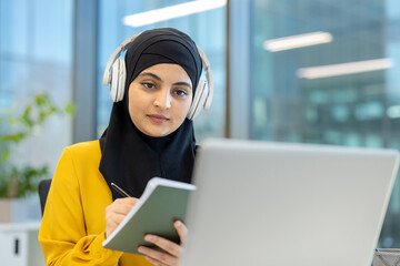 Muslim woman wearing a hijab concentrating while taking notes from a laptop and wearing headphones, engaging in distance learning or professional development in a modern office environment
