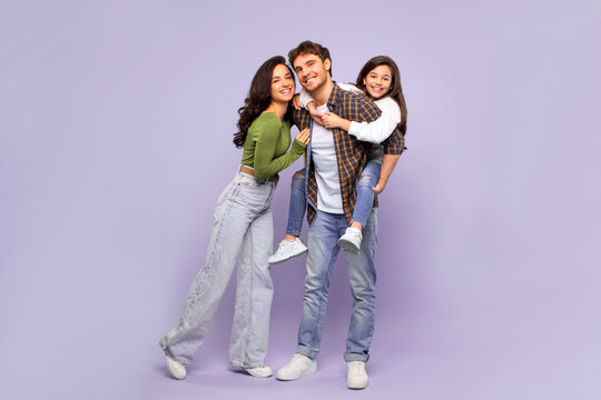 Portrait of happy family of three parents and their daughter having fun over purple studio background, girl piggybacking dad, full length shot
