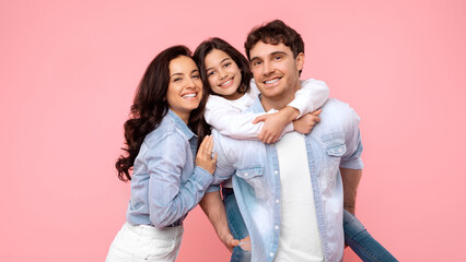 Joyful family, mother dad and daughter, father give piggyback ride to girl, posing together isolated on pink coral background, smiling at camera. Family day concept
