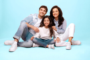 Happy European family sitting on floor and embracing, young parents and their daughter posing on blue studio background, smiling at camera, full length