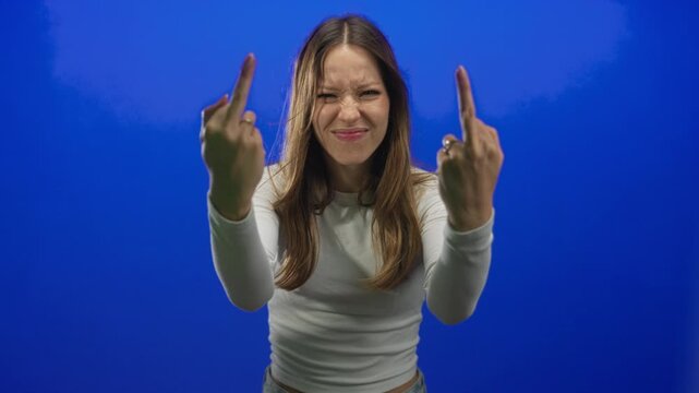 Young hispanic woman with scrunched face showing both middle fingers to camera in bright blue studio; anger defiance.
