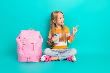Cute girl sitting on teal background holding a smartphone with her finger pointing confidently,...