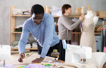Experienced male fashion designer drafting clothing pattern by hand on craft paper at work table in creative studio, while female colleague working on fitting dress on mannequin in background..
