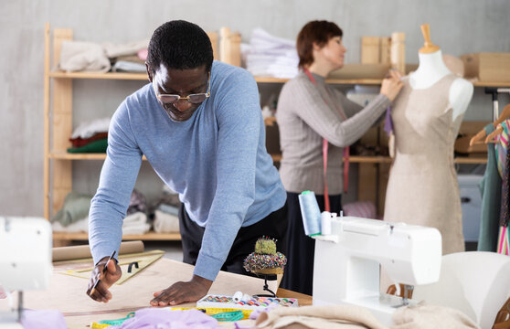 Experienced male fashion designer drafting clothing pattern by hand on craft paper at work table in creative studio, while female colleague working on fitting dress on mannequin in background.. - Powered by Adobe