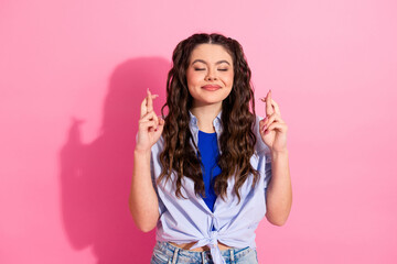 Smiling young lady crossing fingers for good luck in casual outfit standing against a pink...