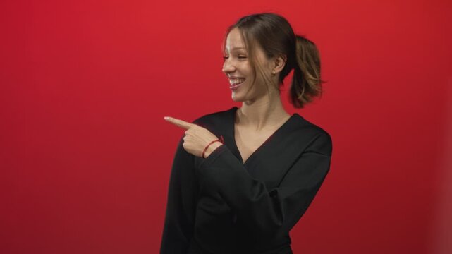Woman pointing finger to the left in red studio wearing a black top and ponytail, smiling broadly; joy playful energy.