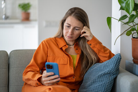 Serene relaxed woman focused on reading or engaging with content on smartphone seated comfortably on home couch. Female calm expression conveys deep concentration, positive leisure engagement - Powered by Adobe