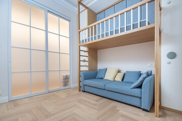 A cozy small apartment features a loft bed above a comfortable blue sofa. Light floods in from frosted glass doors, creating a welcoming and stylish atmosphere.