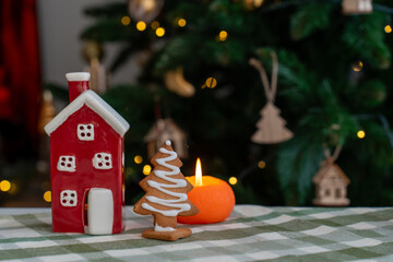 Red ceramic house decoration with gingerbread Christmas tree cookie and burning candle on festive table with bokeh lights. Concept of holiday home decor, cozy winter atmosphere, seasonal celebration.