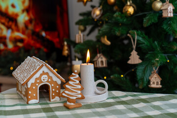 Gingerbread house and Christmas tree cookies with white icing next to burning candle on festive table with decorated tree. Concept of holiday baking traditions, cozy celebration and winter home decor.