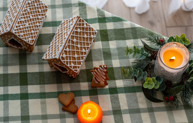 Decorated gingerbread houses with lattice icing pattern, heart cookies and burning candles with evergreen wreath on checkered tablecloth. Cozy home decoration and festive atmosphere.