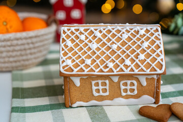 Homemade gingerbread house with white icing decoration on checkered tablecloth, with blurred Christmas background and basket of tangerines. Festive winter holidays and traditional homemade sweets.