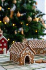 Gingerbread house decorated with white icing on checkered tablecloth in front of Christmas tree with golden lights and ornaments. Concept of winter holidays, homemade sweets and festive atmosphere.