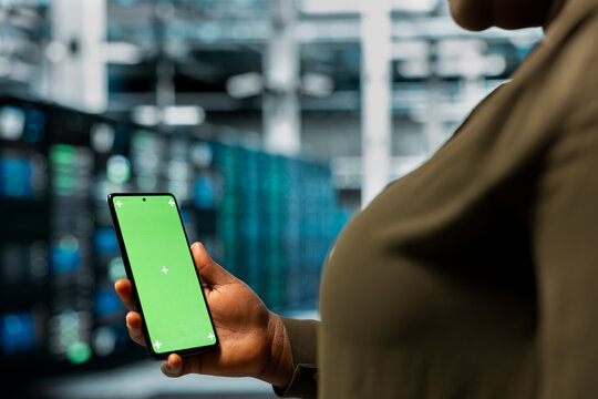 Close up of data center IT specialist using green screen smartphone to monitor mainframes network performance. African american woman in server room checks gear rigs functionality using mockup phone
