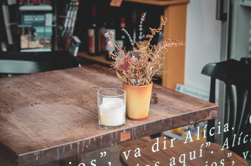 Rustic wooden table near a window with a glass of white beverage and a pot of dried flowers. Through the window, books and bottles are visible, with Spanish text printed on the glass