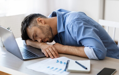 Fototapeta premium Sleep At Work. Exhausted overworked man employee napping at workplace in modern office, tired after working with laptop and reports, sitting at desk, placed head on hands, having job fatigue