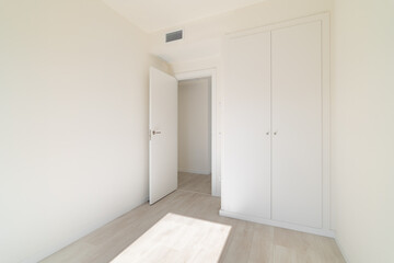 A bright, empty room with white walls and light wooden flooring. A closed door and a closet are visible. Natural light enters through a window.
