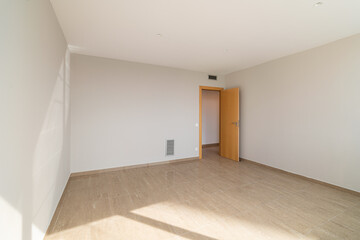 Empty room with light-colored walls and wooden floor. A single door leads to another space. Natural light enters through a window, creating a bright atmosphere.