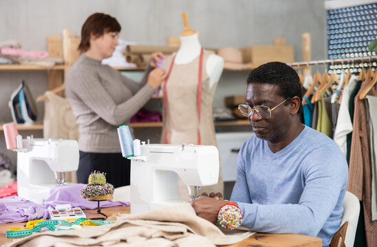 African American man tailor sews clothes in sewing machine. Worker uses sewing machine to create clothes for clients. Companion worker in background with mannequin.