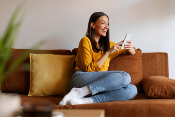 Positive woman sitting on couch using cellphone, smiling looking at screen, enjoying doing online ecommerce shopping or watching videos