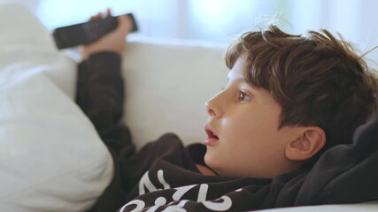 Close-up of an 8-year-old boy sitting on the sofa, fully absorbed in what he’s watching on TV, mouth open in amazement, holding a remote control in his hand.