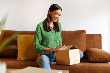 Happy young woman customer receiving online order, opening carboard box, sitting on sofa at her home interior, free space