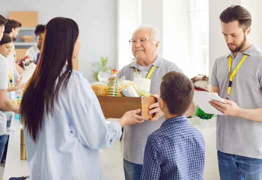 Mature cheerful volunteer working at charity center and giving free food donations for people in need in charitable foundation. Family of mother with child boy receiving humanitarian aid.