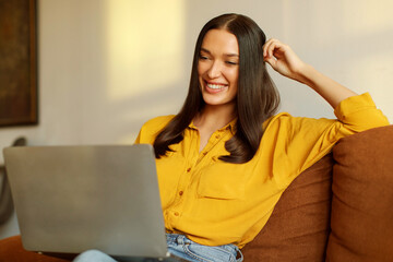 Smiling lady freelancer working from home, sitting on couch and using laptop, surfing on internet...