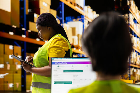 African american staff uses package tracking interface on tablet to check on the delivery service, industrial warehouse filled with storage racks. Efficient online shopping logistics.