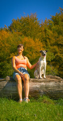 PORTRAIT: Smiling young woman in shorts and orange tank top rests on a fallen log with her obedient doggie beside her in a sunny meadow, surrounded by colorful autumn forest under a clear blue sky.